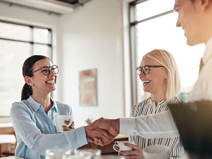 Due donne e un uomo in un ufficio si stringono la mano sorridenti mentre prendono un caffè